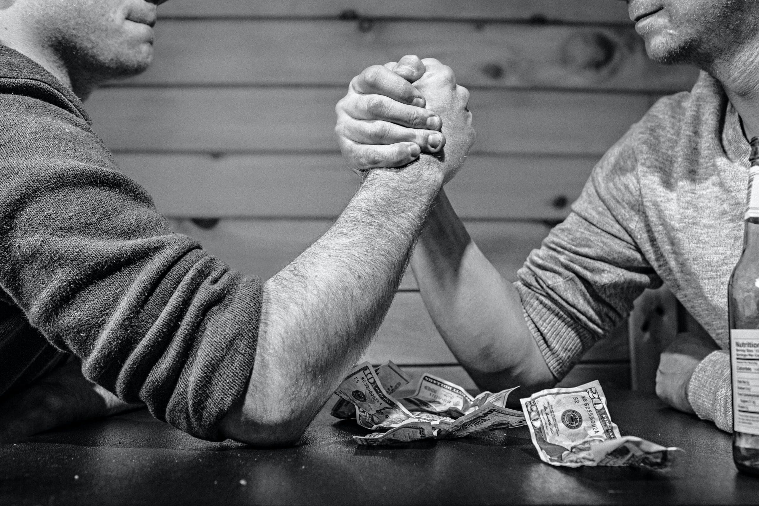 Two men arm wrestling on a table, beside a pile of dollars